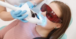 Young woman undergoing professional teeth whitening treatment using a light-activated device, wearing protective red-tinted safety glasses during the procedure.