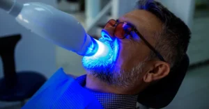 Older man undergoing professional teeth whitening with a blue led light device, wearing protective glasses during in-office cosmetic dental treatment.