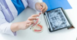 Dentist demonstrating dental procedures using a tooth model and dental tool, with a dental x-ray image and folder visible on the desk in the background.